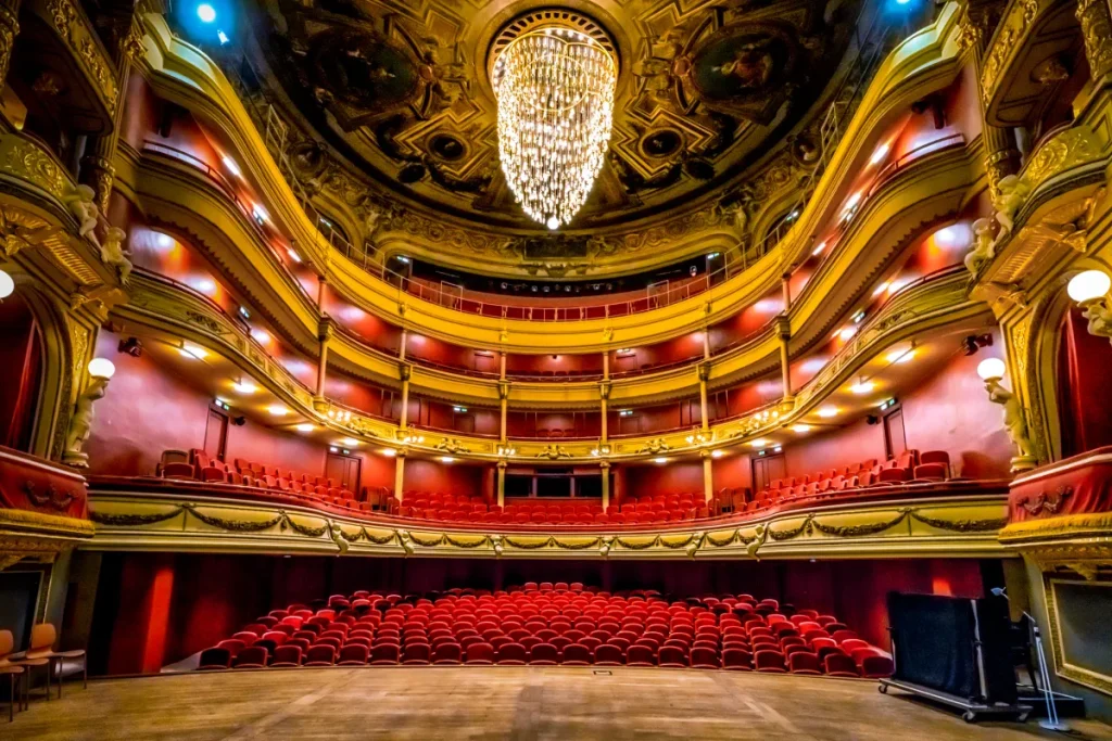 Vue depuis la scène d'une salle d'opéra à l'italienne avec des fauteuils de couleur rouge et un grand lustre lumineux.